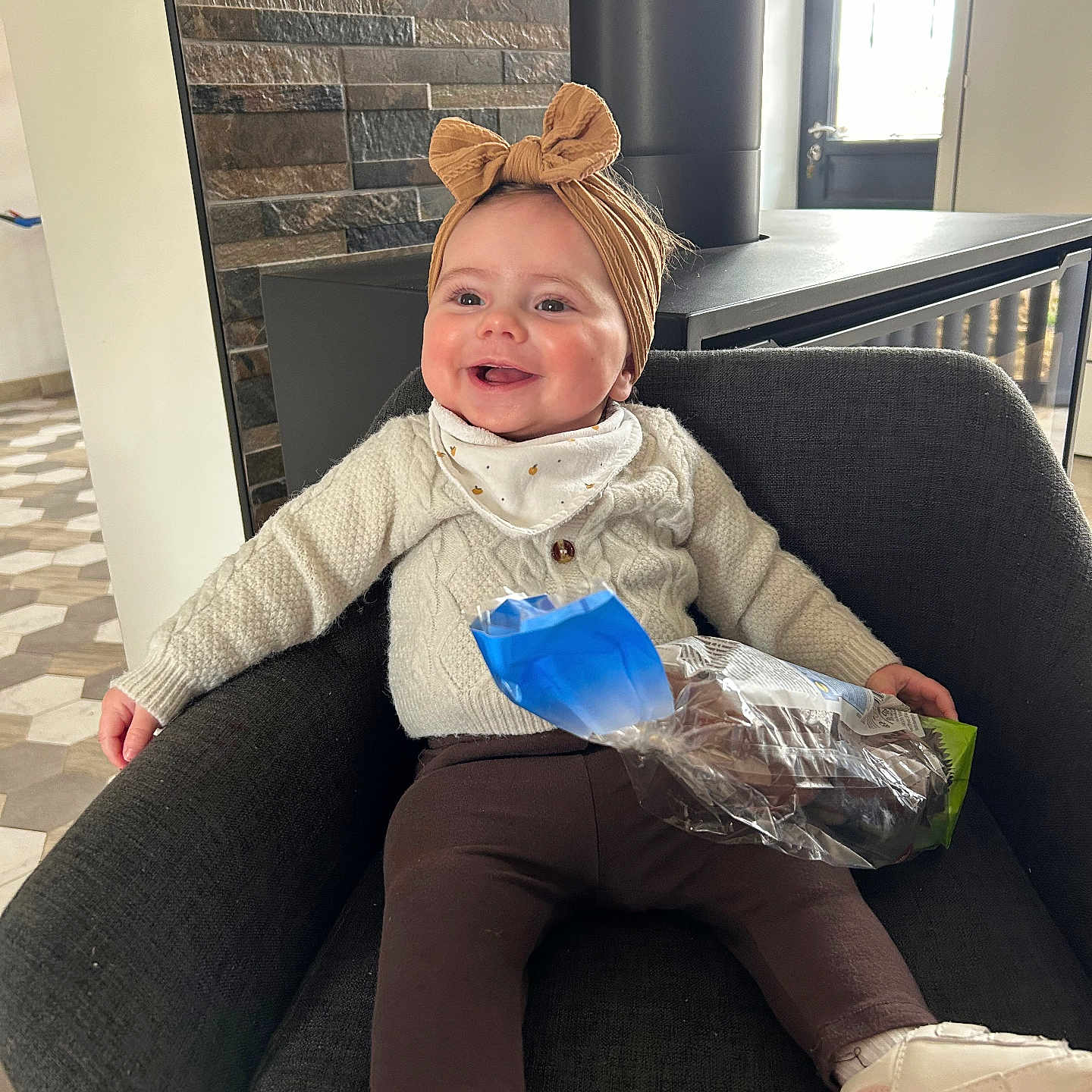 Solyne participe au concours pour gagner de l'argent avec cette photo : baby, child, smiling, headband, knit_sweater, chair, snack_bag, indoor, modern_interior, window, stone_wall, footwear, happy, seated, cozy, person, cute, clothing, portrait, toddler
