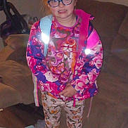 Sarah is registered to the contest to win money with this photo: child, glasses, colorful_clothing, butterfly_pattern, indoor, couch, vacuum_cleaner, carpet, pants, jacket, person, standing, home, furniture, casual, smile, long_hair, female, backpack, room