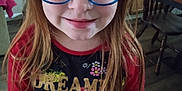 Sarah is registered to the contest to win money with this photo: child, girl, red_hair, glasses, smiling, indoor, kitchen, table, chair, wooden_furniture, long_hair, person, portrait, casual_clothing, floral_pattern, colorful, young, face, eyewear, home