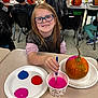 child, glasses, painting, pumpkin, classroom, paintbrush, paint, cup, plate, table, smile, apron, art, kids, school, desk, plastic_apron, orange_pumpkin, creative_activity, happy