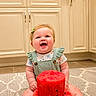 baby, child, infant, smiling, happy, portrait, watermelon, fruit_cake, birthday, cake, sitting, indoor, kitchen, cabinets, rug, green_overalls, floral_shirt, hair_bow, bare_feet, celebration