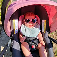 Sage is registered to the contest to win money with this photo: baby, child, sunglasses, heart_shaped, pink, stroller, seatbelt, bib, smile, sunlight, outdoor, sidewalk, grass, happy, infant, person, footwear, shadow, casual, cute