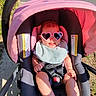 baby, child, sunglasses, heart_shaped, pink, stroller, seatbelt, bib, smile, sunlight, outdoor, sidewalk, grass, happy, infant, person, footwear, shadow, casual, cute