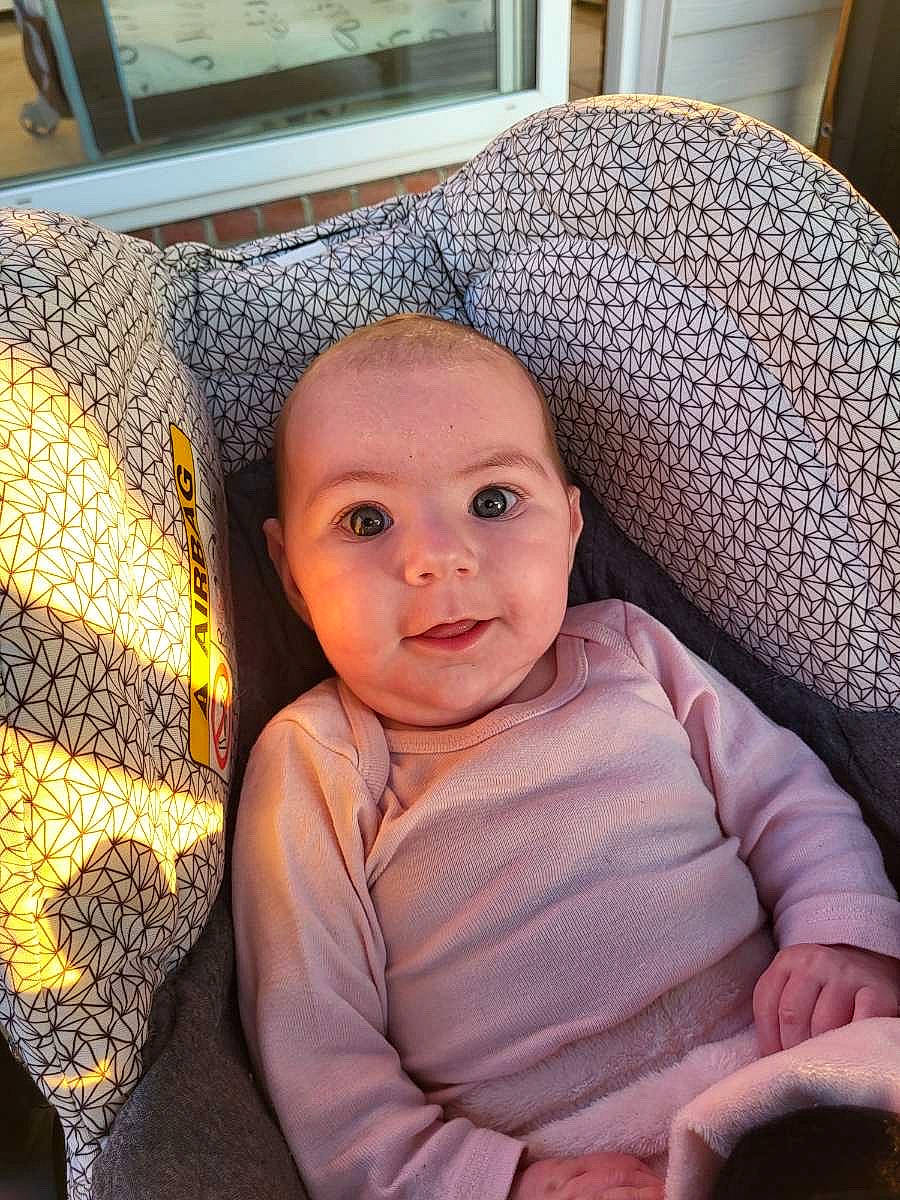 baby, car_seat, child, cozy, cute, face, hand, happy, head, indoors, infant, natural_light, patterned_fabric, pink_clothing, portrait, seat, skin, smiling, window, young_child