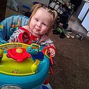 Arianna is registered to the contest to win money with this photo: toddler, child, baby, smiling, sitting, activity_walker, toy, playtime, indoor, home, carpet, chair, colorful, blonde_hair, portrait, face, eyes, clothing, furniture, family_photo