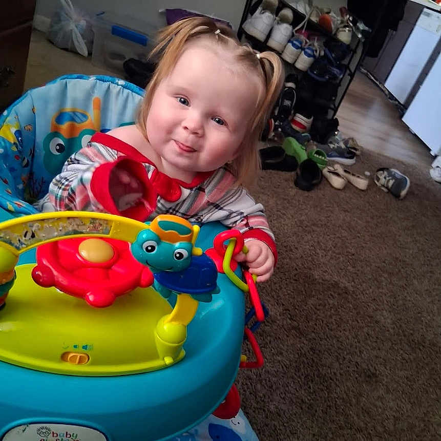 Arianna is registered to the contest to win money with this photo: toddler, child, baby, smiling, sitting, activity_walker, toy, playtime, indoor, home, carpet, chair, colorful, blonde_hair, portrait, face, eyes, clothing, furniture, family_photo