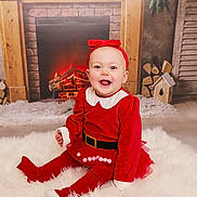 Brooklyn is registered to the contest to win money with this photo: baby, child, red_clothing, headband, smile, happy, indoor, fireplace, holiday, festive, rug, fur_rug, decor, wood, log, toy_house, floor, toddler, portrait, cute