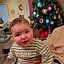 Elliott a rejoint le concours — aidez-le/la à gagner de superbes lots ! baby, child, striped_clothing, face, hand, christmas_tree, holiday_decorations, indoor, person, curious_expression, home, festive, colorful_lights, wood_floor, mirror, furniture, holding, warm_lighting, rings, sweater