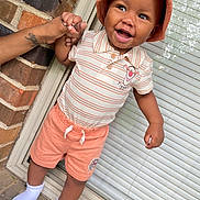 Omari joined the competition — help win amazing prizes! toddler, child, smile, hat, bucket_hat, shorts, striped_shirt, tigger_character, hand_holding, adult_hand, brick_wall, window_blinds, white_socks, outdoor, happy, person, clothing, footwear, portrait, standing