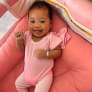 Meadow is registered to the contest to win money with this photo: baby, infant, smiling, pink_clothing, crib, cushion, happy, person, child, cute, lying_down, bracelet, indoor, soft_texture, baby_girl, raised_hand, head, face, relaxing, comfort