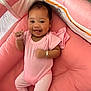 baby, infant, smiling, pink_clothing, crib, cushion, happy, person, child, cute, lying_down, bracelet, indoor, soft_texture, baby_girl, raised_hand, head, face, relaxing, comfort