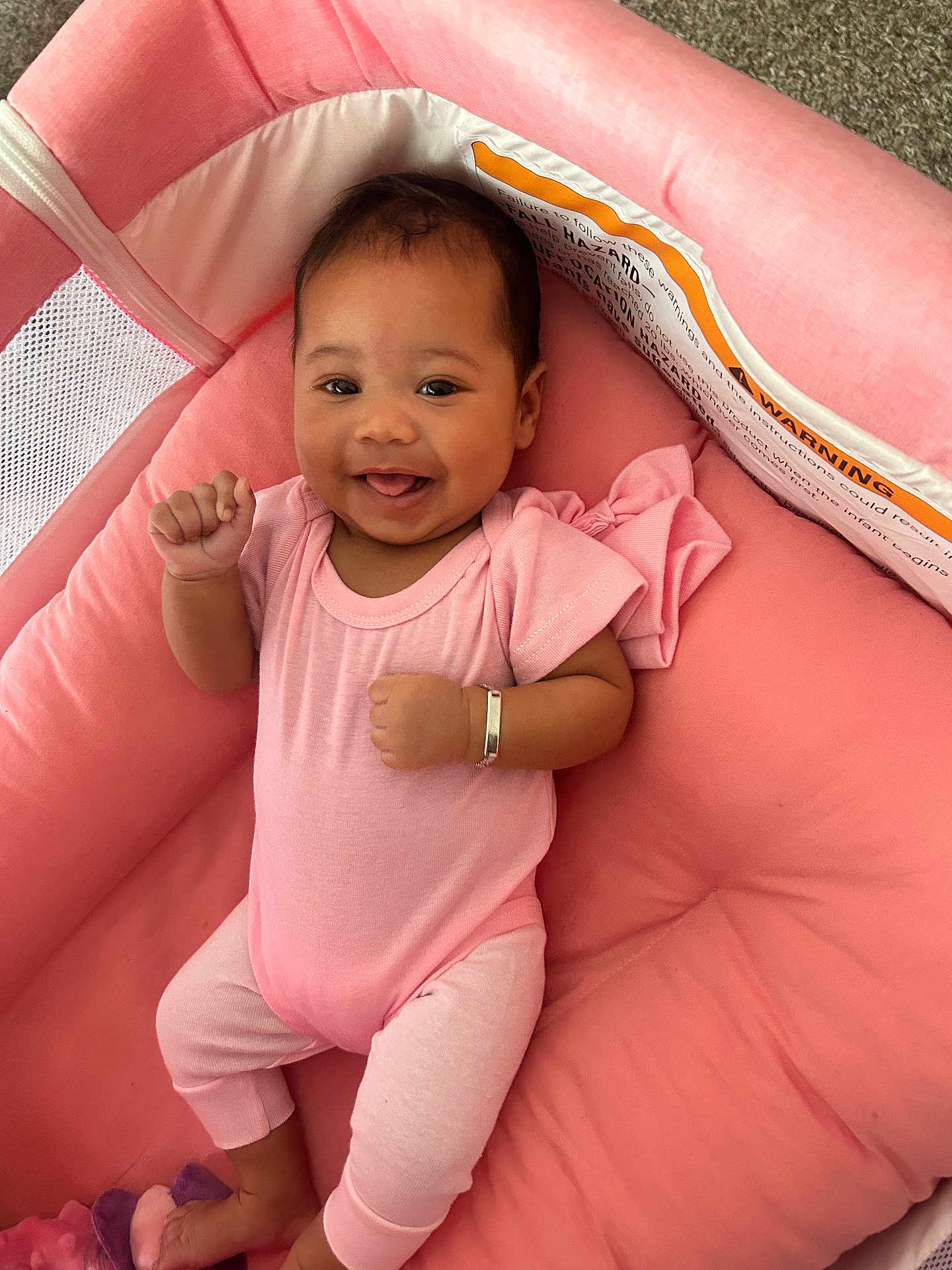 Meadow is registered to the contest to win money with this photo: baby, infant, smiling, pink_clothing, crib, cushion, happy, person, child, cute, lying_down, bracelet, indoor, soft_texture, baby_girl, raised_hand, head, face, relaxing, comfort