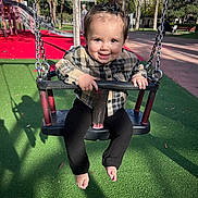 Gabriella a rejoint le concours — aidez-le/la à gagner de superbes lots ! toddler, child, swing, playground, smiling, outdoor, daylight, baby, black_pants, plaid_shirt, barefoot, hair_bow, green_floor, red_floor, metal_chains, palm_trees, shadow, park_bench, person, happy