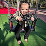 toddler, child, swing, playground, smiling, outdoor, daylight, baby, black_pants, plaid_shirt, barefoot, hair_bow, green_floor, red_floor, metal_chains, palm_trees, shadow, park_bench, person, happy