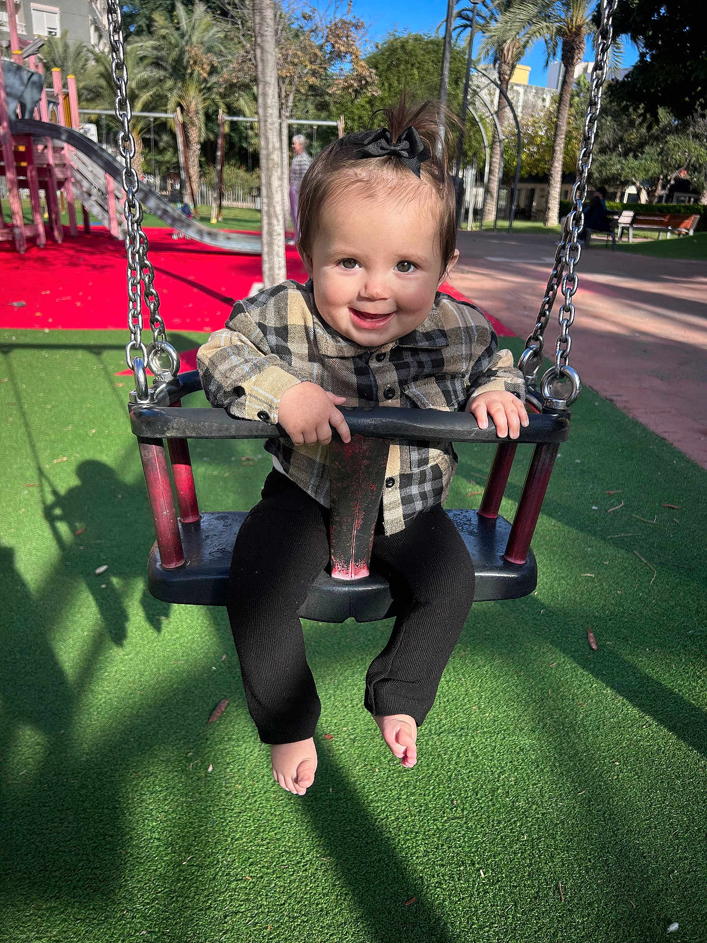 Gabriella a rejoint le concours — aidez-le/la à gagner de superbes lots ! toddler, child, swing, playground, smiling, outdoor, daylight, baby, black_pants, plaid_shirt, barefoot, hair_bow, green_floor, red_floor, metal_chains, palm_trees, shadow, park_bench, person, happy