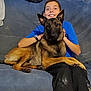 blue_shirt, boy, brown_dog, canine, casual, companionship, couch, cozy, dog, friendship, furniture, happy, human, indoor, lap, pet, relaxed, sitting, smiling, watch