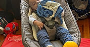 Howard is registered to the contest to win money with this photo: baby, infant, sleeping, bouncer, baby_seat, toy, stuffed_animal, mobile, hanging_toy, blue_clothing, white_socks, safety_harness, indoor, playroom, storage_bins, backpack, wood_floor, colorful, portrait, child