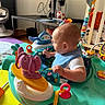 baby, child, infant, activity_center, baby_toys, toy_elephant, playroom, indoor, sitting, side_profile, face, head, bib, onesie, colorful, play_mat, baby_gear, happy, household, portrait
