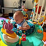 baby, child, infant, activity_walker, playroom, toys, colorful, sitting, smiling, portrait, face, bib, blue_clothing, baby_toys, playmat, crib, basketball, headphones, indoor, happy