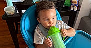 Marcus Nay joined the competition — help win amazing prizes! baby, child, high_chair, sippy_cup, drinking, indoor, kitchen, dining_table, home, portrait, sitting, brown_hair, focused, infant, feeding, blue, green, toy, furniture, household_items