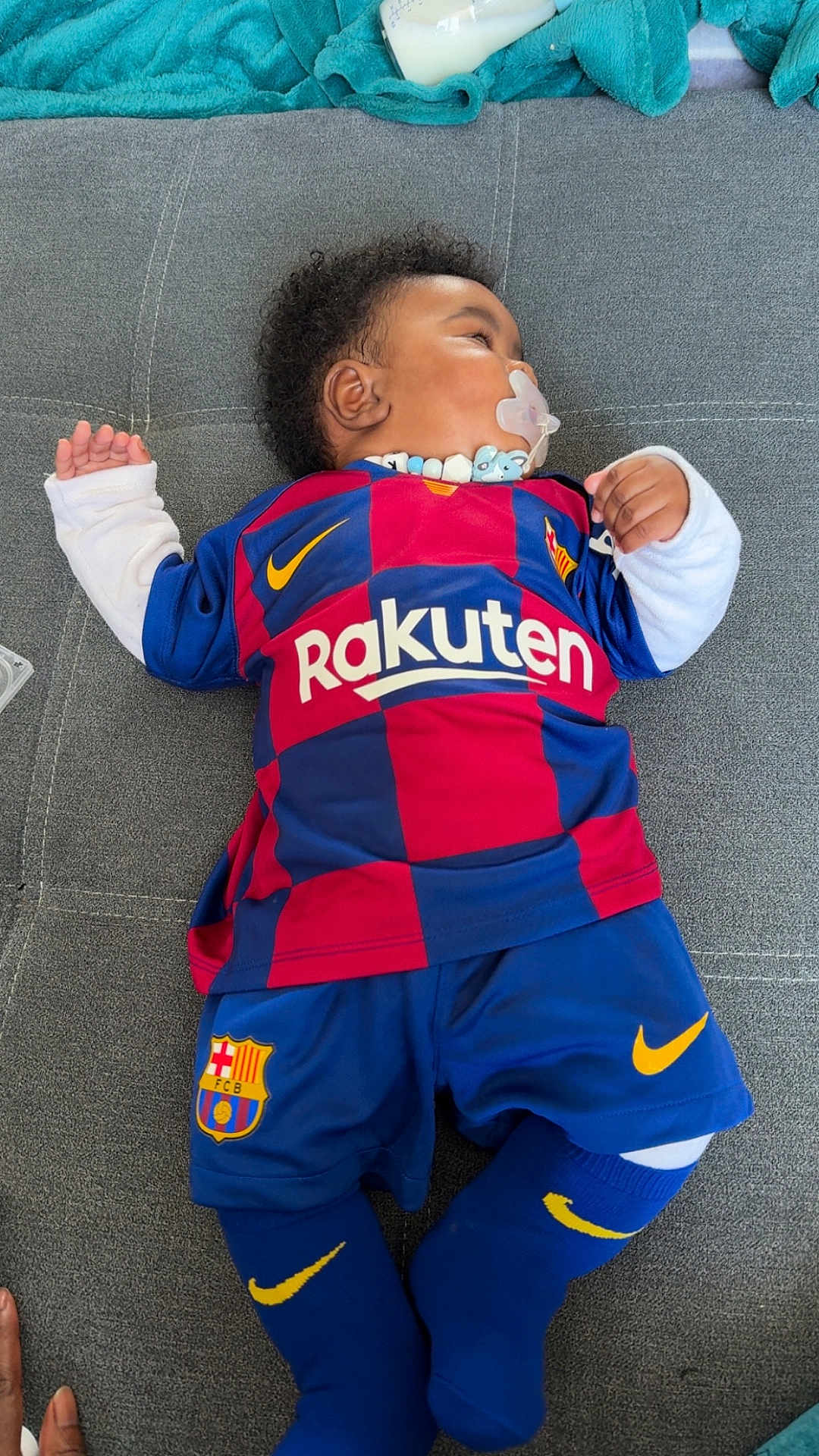 Kaydenn a rejoint le concours — aidez-le/la à gagner de superbes lots ! child, baby, pacifier, pacifier_clip, fc_barcelona_jersey, rakuten_logo, nike_swoosh, sports_shorts, knee_socks, curly_hair, couch, grey_upholstery, teal_blanket, baby_bottle, lying_down, indoor, portrait, infant_clothing, hand, cute