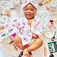 Ruth is registered to the contest to win money with this photo: baby, child, smiling, floral_robe, headband, makeup_brush, cosmetics, lipstick, face, eyelash_curler, beauty_products, sitting, happy, portrait, cute, skin, foot, hand, accessories, white_background