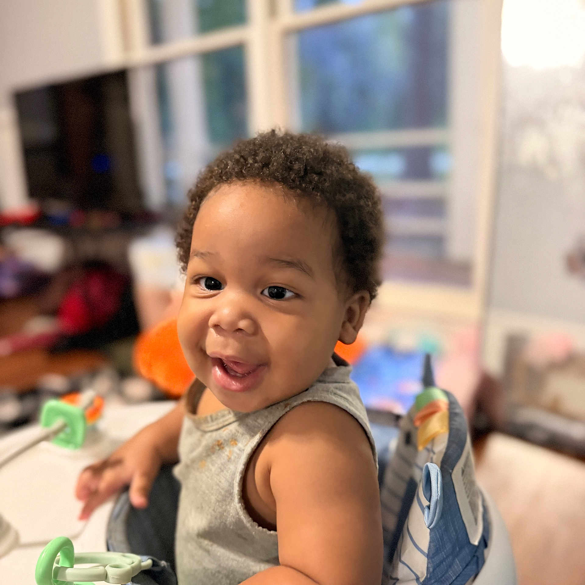 Rammesses joined the competition — help win amazing prizes! baby, child, infant, portrait, smiling, happy, seated, booster_seat, gray_shirt, curly_hair, dark_skin, toy, green_toy, table, indoor, home, window, soft_focus, close_up, candid