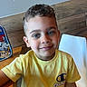 child, boy, smile, yellow_shirt, curly_hair, indoor, wooden_table, backpack, casual_clothing, portrait, happy, seated, blue_eyes, light_skin, chair, wood_paneling, short_sleeves, young, person, casual