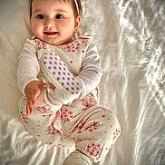Sana participe au concours pour gagner de l'argent avec cette photo : baby, child, infant, pink, headband, shoes, flower_pattern, pajamas, smiling, happy, cute, bed, white_bedspread, playful, portrait, indoors, soft_lighting, clothing, person, lying_down