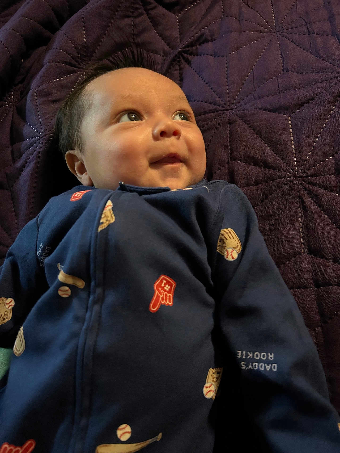 Niklaus is registered to the contest to win money with this photo: baby, infant, face, eyes, smile, onesie, blanket, purple_blanket, lying_down, portrait, close_up, fabric, bedding, pajamas, infant_garment, cute, chubby_cheeks, newborn, indoor, relaxed