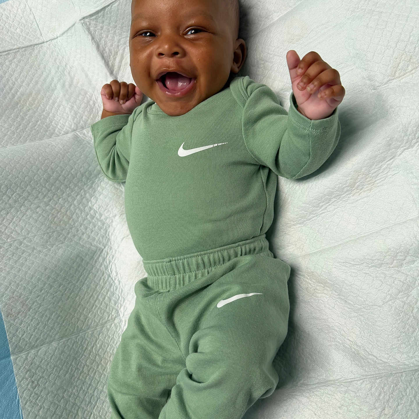 Kyrie Parker is registered to the contest to win money with this photo: baby, smiling, green_clothing, nike_logo, infant, happy, lying_down, textured_surface, white_background, blue_background, small_hands, cute, portrait, child, casual_wear, socks, indoor, baby_garment, joyful, face