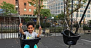 Zayn is registered to the contest to win money with this photo: child, boy, smiling, swing, playground, outdoor, city, urban, park, playground_equipment, seated, swinging, chains, trees, buildings, skyscrapers, fence, pavement, daylight, portrait