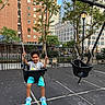 child, boy, smiling, swing, playground, outdoor, city, urban, park, playground_equipment, seated, swinging, chains, trees, buildings, skyscrapers, fence, pavement, daylight, portrait