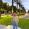 child, boy, smiling, portrait, outdoor, park, palm_trees, boulevard, garden, grass, walkway, street_lamps, evening, outdoor_lighting, bench, jacket, trousers, shoes, landscape, architecture