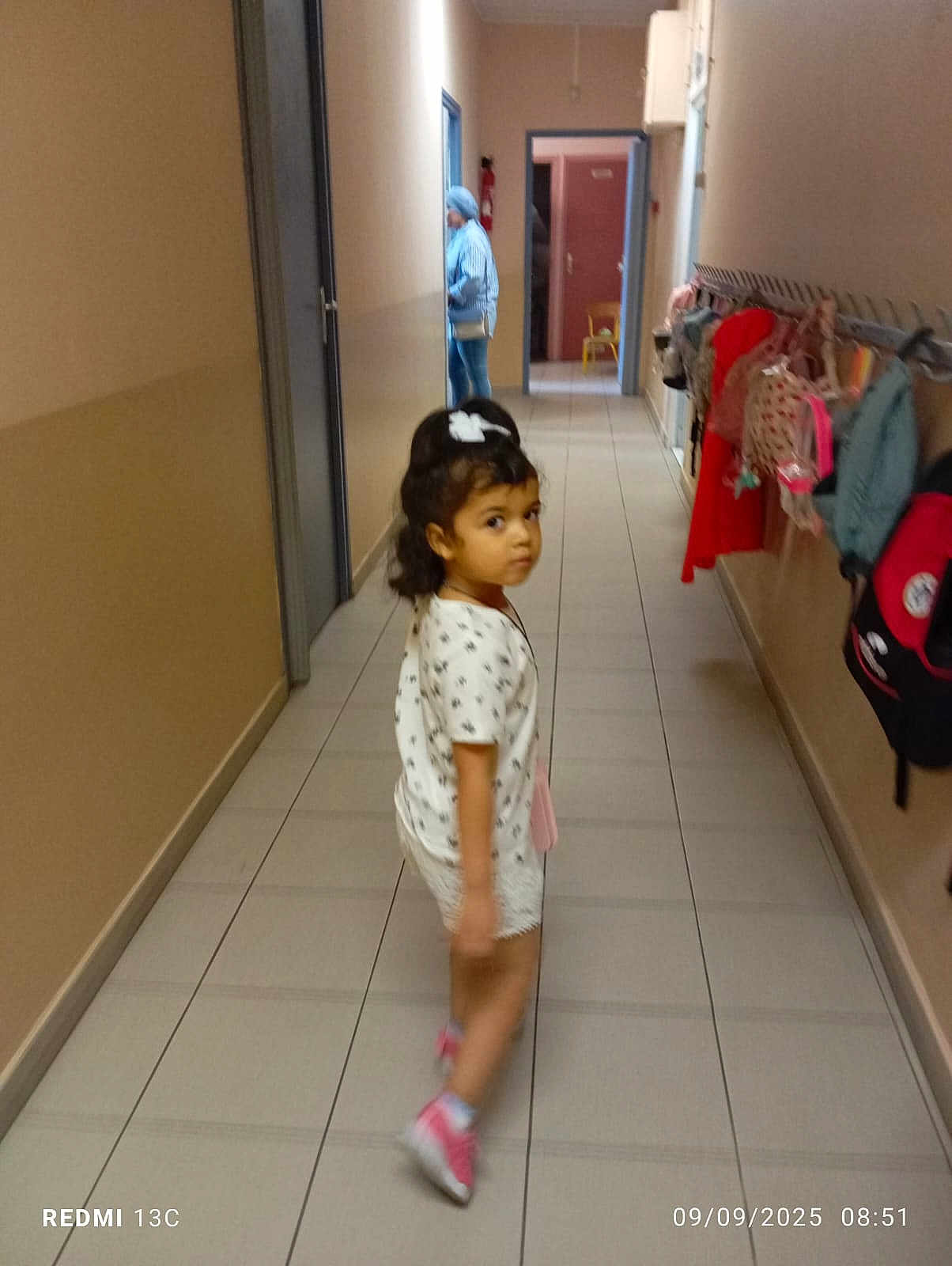 Cilia Soltan a rejoint le concours — aidez-le/la à gagner de superbes lots ! child, girl, hallway, backpacks, walking, tile_floor, white_shirt, shorts, hair_bow, indoor, person, casual_clothing, school, curious, looking_back, footwear, pink_shoes, woman, doorway, bag