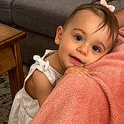 Octavia participe au concours pour gagner de l'argent avec cette photo : toddler, child, baby, girl, pink_bow, white_dress, blanket, couch, wooden_table, carpet, indoor, portrait, cute, smiling, curly_hair, hand, face, eyes, skin, cozy