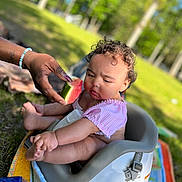 Phorever joined the competition — help win amazing prizes! adult_hand, baby, child, curly_hair, eating, family, feeding, fruit, grass, high_chair, infant, outdoor, park, pink_clothing, portrait, sitting, summer, sunlight, trees, watermelon