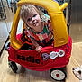 toddler, child, toy_car, red, yellow, indoor, kitchen, wood_floor, dog, pet, curious, colorful_clothing, window, appliances, flooring, playtime, plastic, person, face, vehicle