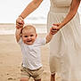 baby, child, beach, sand, walking, adult, hand_holding, white_shirt, shorts, barefoot, smiling, outdoor, summer, parent, woman, dress, happy, first_steps, support, nature