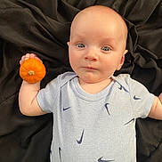 Maverick joined the competition — help win amazing prizes! baby, infant, child, pumpkin, hand, blue_shirt, patterned_clothing, lying_down, black_background, face, skin, small_pumpkin, curious_expression, light_skin, short_sleeves, indoors, person, young_child, holding_object, portrait