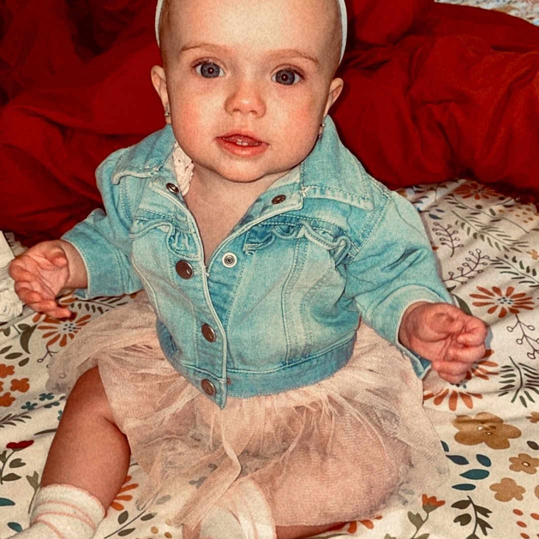 Kambri is registered to the contest to win money with this photo: baby, bedspread, bow, brick, child, cozy, cute, denim_jacket, floral_pattern, headband, indoor, infant, pillow, portrait, seated, smiling, socks, tutu, wall, wood