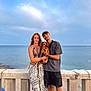 couple, dog, beach, ocean, sky, stone_railing, woman, man, pet, summer, casual_clothing, outdoor, smile, standing, sand, landscape, accessories, hat, bracelet, peaceful