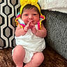 baby, infant, yellow_headband, white_clothing, smile, hands, feet, couch, pillow, patterned_pillow, brown_blanket, indoor, cute, child, portrait, person, happy, headwear, newborn, soft_lighting
