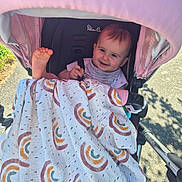 Amelia is registered to the contest to win money with this photo: baby, blanket, canopy, child, clothing, fabric, foot, happy, infant, nature, outdoor, pattern, person, seat, shade, smile, stroller, sunlight, toy, walkway