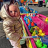 toddler, child, puffer_jacket, gold_jacket, plastic_ducks, fishing_game, net, fairground, outdoor, people, colorful, toy, play, water, game, crowd, casual_clothing, daylight, curious, standing