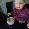 toddler, child, blue_eyes, cereal, bowl, spoon, high_chair, maroon_jacket, indoor, furniture, table, face, person, young_child, meal, snack, expression, hair, seated, cute