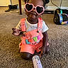toddler, child, sunglasses, heart_shaped, pink_overalls, love_text, baby_bottle, carpet, indoor, cute, fashion, accessories, hair_buns, earrings, playful, smiling, person, casual, home, toy_car