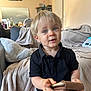 black_shirt, blanket, blonde_hair, blue_eyes, casual, child, couch, curious, expression, furniture, home, indoor, living_room, person, pillow, portrait, soft_light, toddler, wallet, young_child