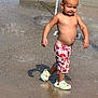 child, toddler, water, splash_pad, summer, bare_chest, shorts, crocs, water_droplets, outdoor, play, fun, sunny, daytime, concrete, grass, happy, walking, boy, casual