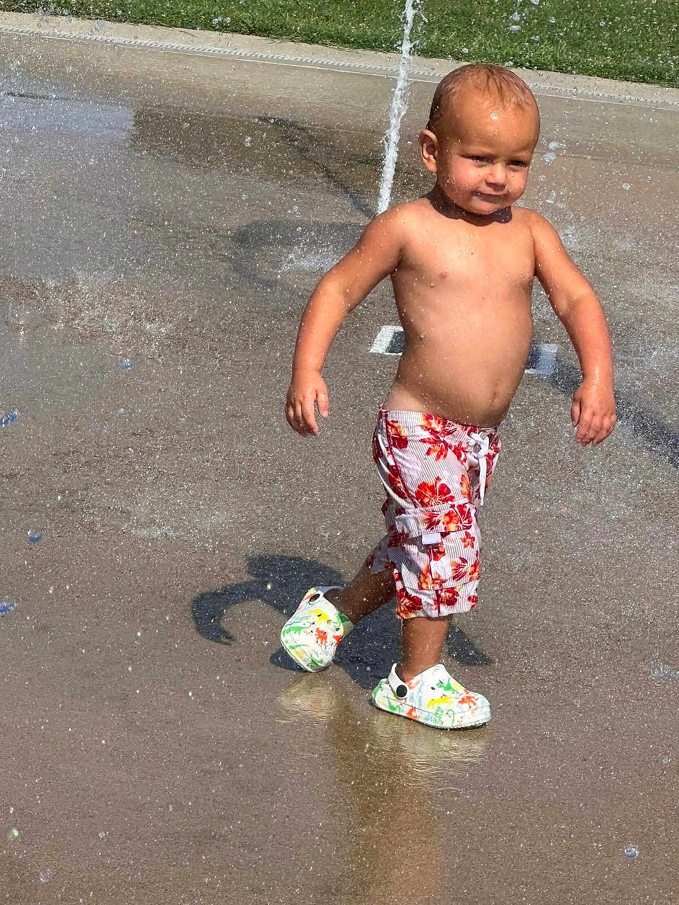 Matteo is registered to the contest to win money with this photo: child, toddler, water, splash_pad, summer, bare_chest, shorts, crocs, water_droplets, outdoor, play, fun, sunny, daytime, concrete, grass, happy, walking, boy, casual